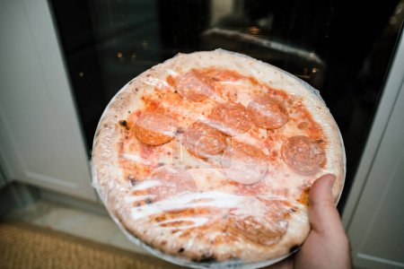 Photo for POV male hand holding deep frozen pizza with salami in hand - covered in plastic film before putting it to the oven - Royalty Free Image