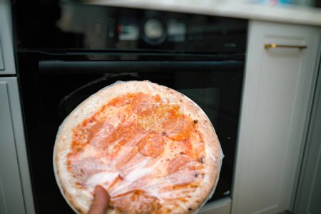 Photo for In fropnt of oven - hand holding deep frozen pizza with salami in hand - covered in plastic film before putting it to the oven - Royalty Free Image