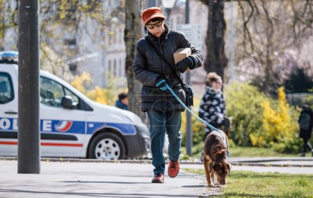 Téléchargez les photos : Strasbourg, France - 29 mars 2023 : Une femme promène son animal de compagnie canin sous le soleil, avec une voiture de police en arrière-plan - en image libre de droit