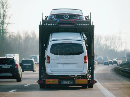 Foto de Alemania - 10 de febrero de 2023: Camión rápido remolque de camión largo blanco que transporta automóviles nuevos por Mercedes-Benz Vito y GL SuV y monta en la autopista de la autopista a la luz del sol bajo el cielo azul - Imagen libre de derechos