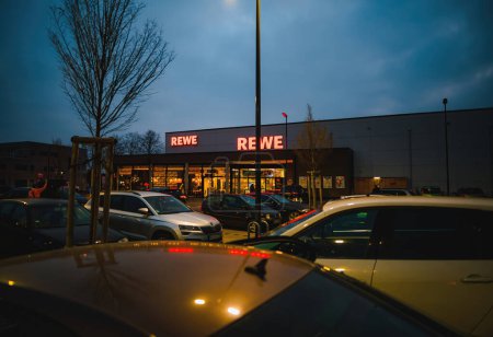Foto de Frankfurt, Alemania - 4 de marzo de 2023: Un supermercado iluminado de REWE por la noche, un símbolo del comercio alemán entre automóviles en una ciudad urbana. - Imagen libre de derechos