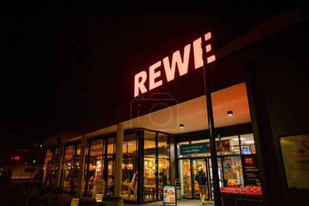 Foto de Frankfurt, Alemania - 4 de marzo de 2023: Supermercado iluminado Rewe en una bulliciosa ciudad alemana, adornado con el logotipo de la compañía y el letrero de neón. Un destino popular para comestibles y otros artículos. - Imagen libre de derechos