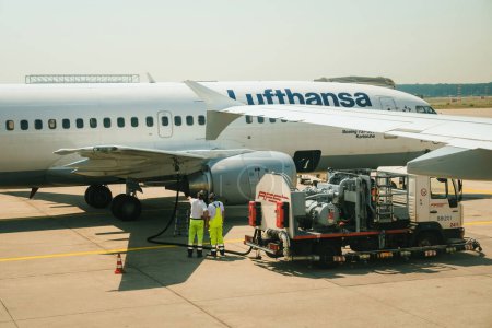 Téléchargez les photos : Allemagne - 6 juil. 2015 : Un Boeing 737-MSN25149 de Lufthansa arrive à un aéroport, entouré de travailleurs en équipement de protection ravitaillant l'avion à réaction en kérosène pour son prochain départ. - en image libre de droit