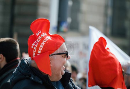 Foto de Strasborg, Francia - 29 de marzo de 2023: Un anciano sonriente se encuentra entre una multitud de hombres adultos en una protesta francesa, llevando un sombrero con un mensaje importante al gobierno sobre el aumento de la reforma de la edad de jubilación. - Imagen libre de derechos