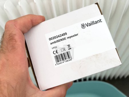 Photo for London, United Kingdom - Jan 1, 2022: A close-up of a male hand holding a new Vaillant Ambisense Repeater VR 52, connected to the internet and near a radiator. A heat pump IoT device. - Royalty Free Image