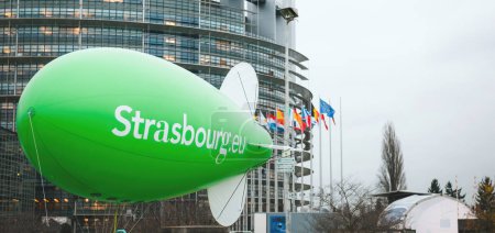 Téléchargez les photos : Les gens travaillent à préparer un grand ballon écologique dirigeable vert avec Strasbourg en grosses lettres, sur fond d'édifices du Parlement. - en image libre de droit