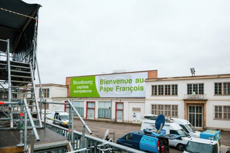 Téléchargez les photos : Strasbourg, France - 24 nov. 2014 : Une bannière massive est drapée sur un bâtiment, accueillant le pape François en français. De nombreux camions des médias diffusent l'événement en direct de la scène. - en image libre de droit