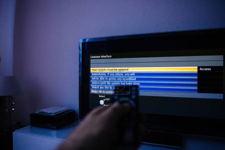 Photo for A man holds a remote while viewing a modern television displaying the message, Your PCMCIA module must be updated immediately to access the service of air and satellite signals. Press OK to continue - Royalty Free Image