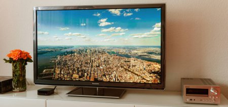Photo for PARIS, FRANCE - NOV 21, 2015: An Apple TV 4K device sits next to a Panasonic Plasma OLED display, projecting an aerial city screensaver. Technology indoors next to onkyo hi-fi device - Royalty Free Image