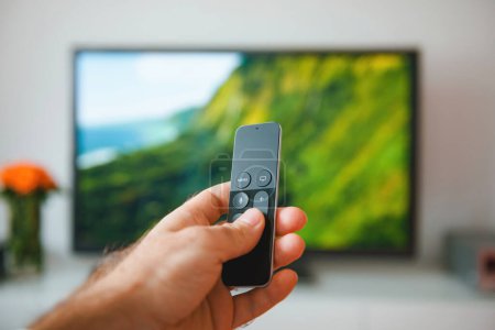 Photo for A young adult lies comfortably in their home, holding a remote control and turning on the television set. They focus on modern technology as they enjoy the cozy domestic life of watching TV indoors - Royalty Free Image