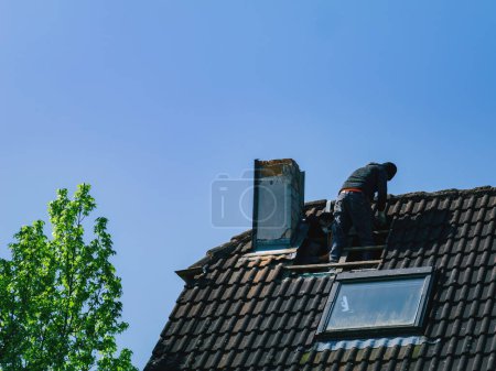 Foto de Un techador se levanta sobre una casa alta, demoliendo la chimenea mientras el cielo azul claro se extiende sobre ellos. La arquitectura de los edificios y los techos de azulejos siguen siendo majestuosos bajo sus pies. - Imagen libre de derechos