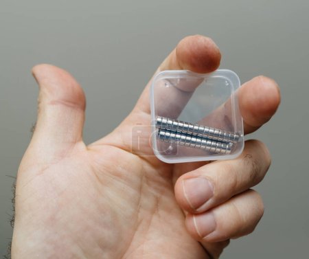 Photo for A single hand holds up a transparent package with multiple round magnets The fingers grip is tight and precise against gray background - Royalty Free Image