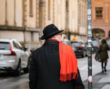 Téléchargez les photos : Strasbourg, France - 12 avril 2023 : Un adulte à la mode portant un chapeau et une écharpe rouge dans une rue animée de la ville avec une architecture, des voitures et des transports remarquables qui incarnent une vie urbaine animée. - en image libre de droit