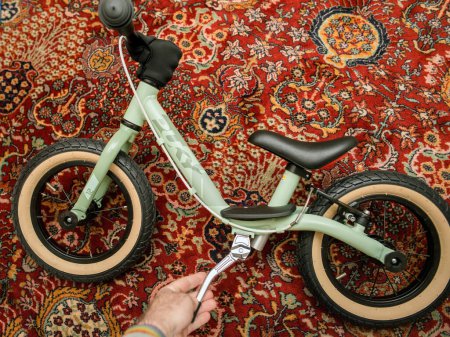 Foto de La mano adulta prueba la nueva bicicleta de tamaño infantil en una alfombra de salón. Listo para la aptitud y el transporte saludable, listo para viajar. Vista de ángulo alto. - Imagen libre de derechos