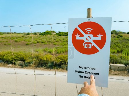 Photo for Male hand pointing to warning sign in red text with symbols against clear sky - no drones - Restricted area with no fly zone and security. Fence, road, plants in rural scene - Royalty Free Image