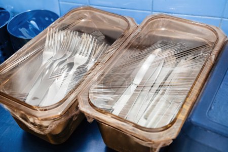 Foto de Cajas de plástico con tenedores y cuchillos en la mesa de comedor de acero inoxidable - cubiertas con película de plástico limpia - Imagen libre de derechos