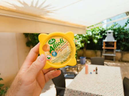 Photo for Mallorca, Spain - Jun 30, 2023: A hand holding Chovi brand aioli sauce in yellow plastick package on an outdoor terrace table. Mediterranean flavors in a kitchen setting. - Royalty Free Image