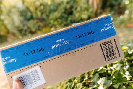 Téléchargez les photos : Hambourg, Allemagne - 24 juil. 2023 : Main masculine POV tenant une nouvelle parcelle de carton Amazon Prime avec fond vert jardin et scoth tape bleue mettant en vedette Prime Day 11-23 juillet - en image libre de droit