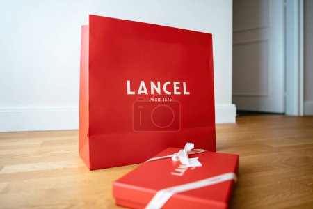 Foto de París, Francia - 25 / 08 / 2023: Vista lateral de la caja de regalo roja de la marca Lancel Paris con elegante logotipo y cinta de seda en el suelo de madera. Un símbolo de lujo y sofisticación. Perfecto para presentaciones - Imagen libre de derechos