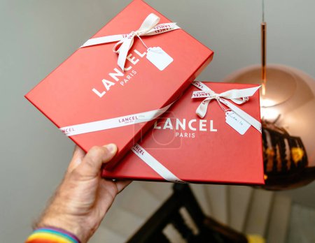 Téléchargez les photos : Paris, France - 25 août 2023 : Homme tenant dans un magasin de luxe deux articles de mode chers Lancel Paris cadeaux en paquets rouges - en image libre de droit