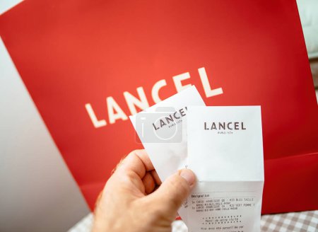 Foto de París, Francia - 25 / 08 / 2023: POV mano masculina sosteniendo el recibo de la tienda de casa de moda Lancel con bolsa de regalo con logotipo en el fondo - Imagen libre de derechos