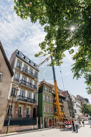 Téléchargez les photos : Strasbourg, France - 7 juil. 2023 : Une grue jaune vibrante se dresse au milieu d'une rue piétonne, où des travailleurs dévoués restaurent une charmante maison alsacienne, préservant les régions riches en immobilier - en image libre de droit