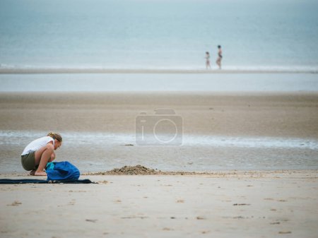 Téléchargez les photos : Neufchatel-Hardelot, France - 18 août 2023 : Une fille profite de moments ludiques dans le sable à Hardelot Beach, avec une silhouette déconcentrée de touristes en arrière-plan - en image libre de droit