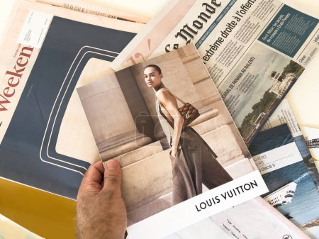 Foto de París, Francia - 18 de septiembre de 2023: Una mano masculina se muestra agarrando una revista que cuenta con publicidad trasera para Louis Vuitton. En el fondo, las ediciones de Le Monde y los periódicos del Financial Times son - Imagen libre de derechos