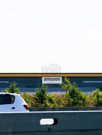 Téléchargez les photos : France - 17 août 2023 : Une voiture blanche circule le long d'une autoroute adjacente à un entrepôt amazonien, qui arbore un logo bien en vue sur sa façade. - en image libre de droit