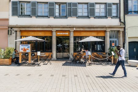 Téléchargez les photos : Strasbourg, France - 7 juil. 2023 : Par une journée ensoleillée d'été, un homme noir se promène délibérément devant le restaurant BioBurger situé à Strasbourg - en image libre de droit