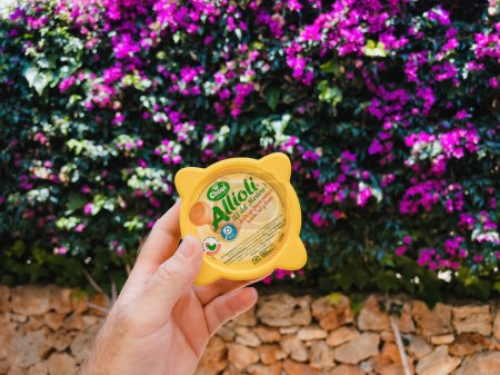 Photo for Mallorca, Spain - Jun 30, 2023: A hand holds a bottle of Chovi Aioli sauce, positioned against a vibrant backdrop of Bougainvillea flowers. - Royalty Free Image
