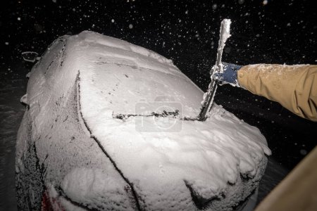 Foto de En medio de una tormenta de nieve, una mano de hombre se esfuerza por eliminar el hielo y la nieve de los limpiaparabrisas de un automóvil fuertemente cubierto. - Imagen libre de derechos