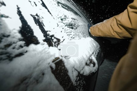 Foto de Capturado contra la oscuridad nocturna, una mano masculina en un guante retira cuidadosamente la nieve de las ventanas congeladas de un automóvil después de una tormenta severa - Imagen libre de derechos