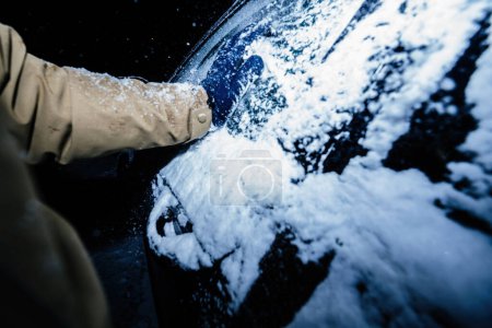 Téléchargez les photos : Capturé tôt le matin dans l'obscurité, un homme enfermé dans un gant nettoie avec diligence la neige d'une voiture gelée à la suite d'une tempête. - en image libre de droit