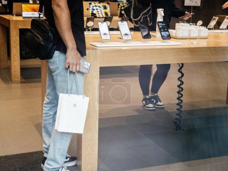 Téléchargez les photos : Paris, France - 22 sept. 2023 : Un client tient un sac en papier contenant le nouvel iPhone 15 Pro, debout à côté de la gamme complète des derniers modèles d'iPhone affichés dans le magasin. - en image libre de droit