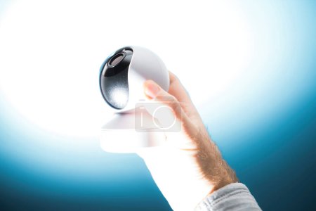 Foto de Una mano masculina sostiene una cámara de vigilancia dirigida hacia arriba, colocada sobre un fondo azul-blanco, que ilustra la tensión entre la tecnología y la privacidad personal. - Imagen libre de derechos