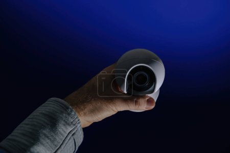 Foto de Una mano masculina sostiene una cámara de vigilancia sobre un fondo azul, simbolizando la interrupción de la privacidad y la intrusión - Imagen libre de derechos
