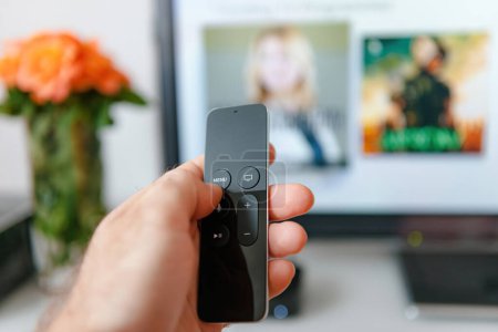 Photo for Paris, France - Nov 25, 2015: Mans hand holds an Apple TV 4K remote, expertly navigating with the menu button, against a large, defocused television screen in the background. - Royalty Free Image
