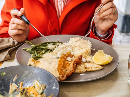 Foto de Mujer en un abrigo rojo hábilmente usando tenedor y cuchillo para saborear un delicioso plato de pescado, complementado con limón y arroz, en una terraza al aire libre de un restaurante - Imagen libre de derechos
