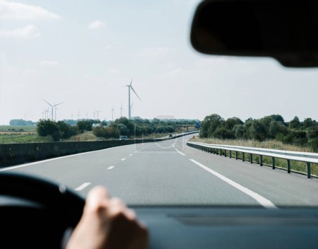Téléchargez les photos : Un plan concentré d'une femme main sur le volant alors qu'elle conduit sur une autoroute française peu peuplée avec une ligne de moulins à vent générateurs d'électricité au loin - en image libre de droit