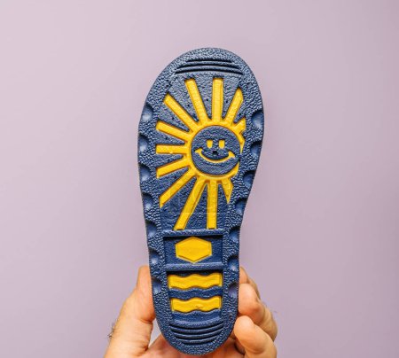 Téléchargez les photos : Prise de vue d'une main masculine tenant des bottes en caoutchouc pour enfants ornées de marques jaunes et d'un soleil souriant, sur fond gris - en image libre de droit