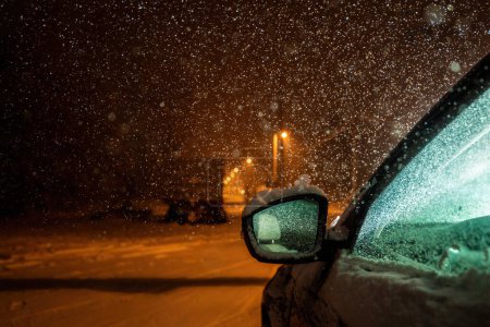 Téléchargez les photos : Image réelle convaincante : Voiture éclairée de l'intérieur, passager arrière vide réfléchi dans le rétroviseur, et une route vide éclairée par un réverbère en perspective lors d'une tempête hivernale - en image libre de droit