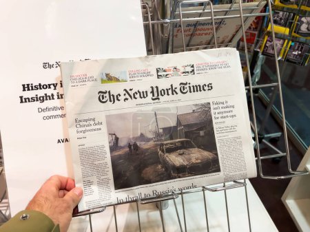 Foto de Londres, Reino Unido - 21 abr 2023: mano alcanza el último número del periódico The New York Times en un quiosco de prensa, con una imagen de portada de un pueblo bombardeado en Ucrania - Imagen libre de derechos