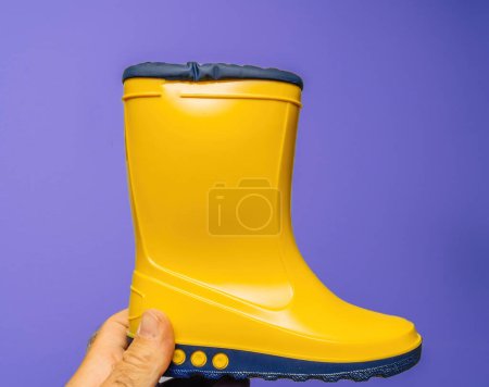 Photo for POV: Male hand presents vibrant yellow childrens boots against a white and violet backdrop - Royalty Free Image