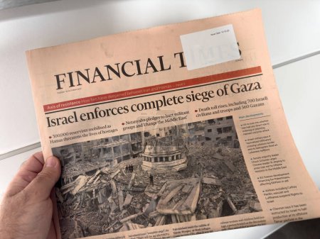 Photo for Paris, France - Oct 10, 2023: POV: A male hand carefully reads the latest Financial Times newspaper with the headline Israel Enforces Complete Siege of Gaza, featuring a picture of a war-torn urban - Royalty Free Image