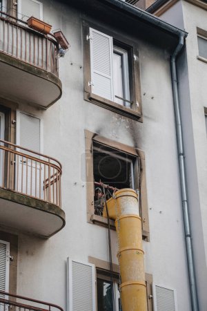 Téléchargez les photos : Strasbourg, France - 22 nov. 2023 : Un travailleur près d'un seau d'ascenseur jaune près d'une fenêtre d'un bâtiment urbain, nettoyant les débris après un incendie à l'intérieur - en image libre de droit