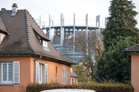 Foto de Destacan las casas naranjas tradicionales con techos de azulejos, con el imponente Parlamento Europeo en Estrasburgo visible en el fondo en medio de los árboles. - Imagen libre de derechos