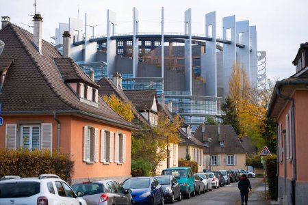 Foto de Estrasburgo, Francia - 22 de noviembre de 2023: Destacan las casas naranjas tradicionales con techos de azulejos, con el imponente Parlamento Europeo en Estrasburgo al fondo: peatones y coches - Imagen libre de derechos