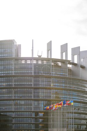 Téléchargez les photos : Une vue ensoleillée du bâtiment du Parlement européen à Strasbourg, avec son architecture moderne distinctive, accentuée par de multiples drapeaux nationaux flottant au premier plan. - en image libre de droit