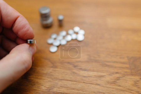 Description: A focused shot of fingers pinching a single button battery, with an array of batteries blurred in the background on a wooden surface